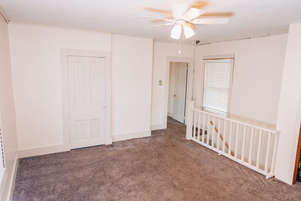 Upstairs bedroom at 11 Sherman Street, Dennison, Ohio featuring ceiling fan, window with blinds, closet door, and railing overlooking staircase.